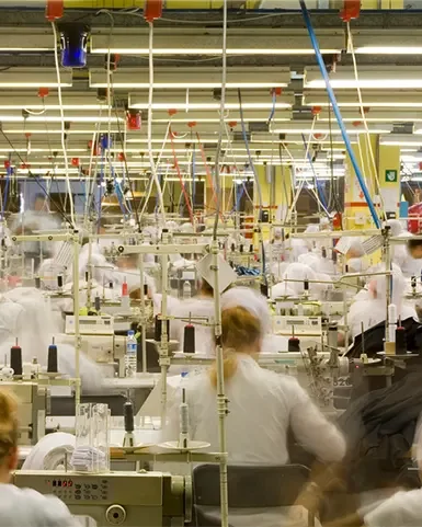 Viele Menschen, vermutlich Frauen, sitzen an Nähmaschinen in einer großen, hell beleuchteten Näherei. Das Foto ist etwas unscharf, wodurch  eine gewisse Schnelligkeit zu erahnen ist.